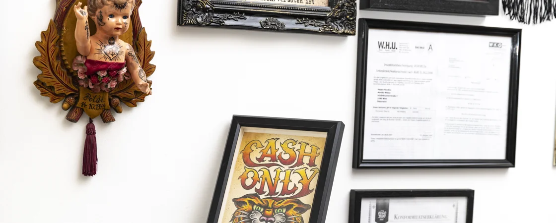 Das Bild zeigt eine Wand, die mit verschiedenen gerahmten Gegenständen dekoriert ist. Auf der linken Seite befindet sich die Figur eines Babys mit Tattoos auf einem verzierten Holzschild. Rechts davon sind mehrere gerahmte Bilder und Dokumente an der Wand befestigt.