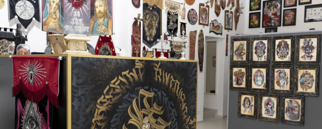 FAQ | Das Bild zeigt das Innere eines Tattoo-Studios oder einer Kunstgalerie mit einer beeindruckenden Sammlung von Tattoo-Designs und Kunstwerken. Die Wände sind vollständig mit gerahmten Tattoo-Vorlagen bedeckt, die verschiedene Motive wie Totenköpfe, Porträts, traditionelle Tattoo-Designs und künstlerische Illustrationen zeigen. Im Zentrum des Raumes befindet sich ein großes, dunkles Kunstwerk mit goldenen Verzierungen und Text. Links im Bild sind rote Banner oder Fahnen mit religiösen oder mystischen Symbolen zu sehen.