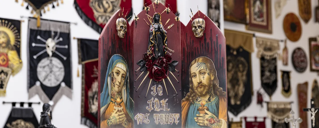 Instagram-Feed | Das Bild zeigt ein dekoriertes religiöses Objekt mit Jesus und Maria mit Tattoos, das vor einer Wand voller religiöser Banner und anderer Objekte steht. Das Objekt ist dreiteilig und hat eine dunkle, rote Farbe. Auf der linken Seite befindet sich ein Bild von Maria mit Tattoos und Heiligenschein. Auf der rechten Seite befindet sich ein Bild von Jesus mit Tattoos und Heiligenschein. In der Mitte steht eine dunkle Madonnenfigur, umgeben von dunklen Rosen. Über den Bildern von Maria und Jesus befinden sich Schädel. Die Inschrift "In Ink We Trust" ist auf der Tafel zu lesen. Der Hintergrund ist voll von religiösen Bannern und gerahmten Bildern.