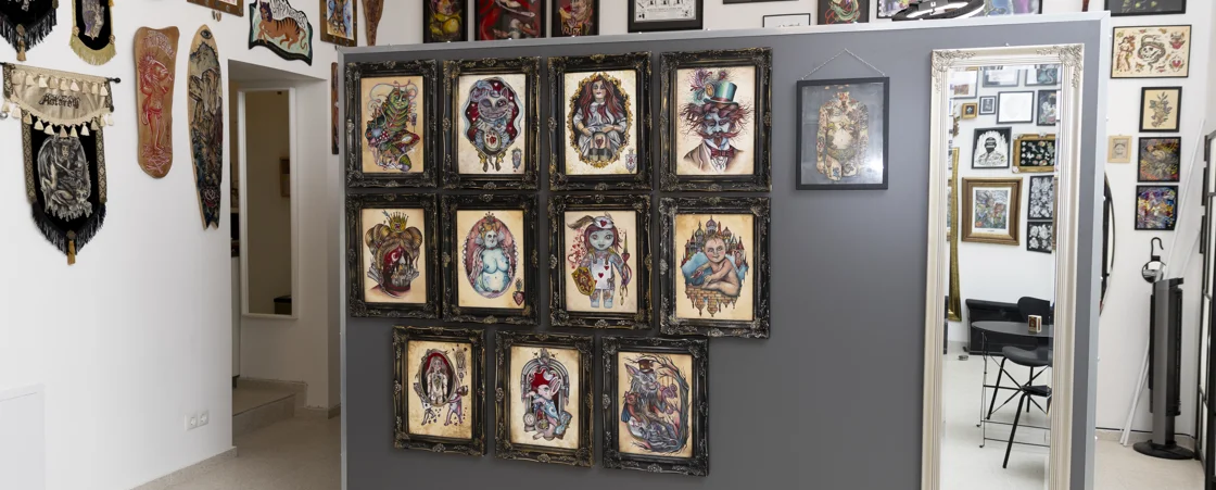 Monikas Bilder | Das Bild zeigt das Innere des Tattoo-Studios von Happy Needles. Die Wände sind mit einer Vielzahl von Kunstwerken geschmückt, darunter gerahmte Illustrationen, Wandteppiche und bemalte Holzbretter. Ein großer Spiegel an der rechten Seite des Bildes spiegelt einen weiteren Raum wider, der mit weiteren Kunstwerken und Möbeln gefüllt ist.