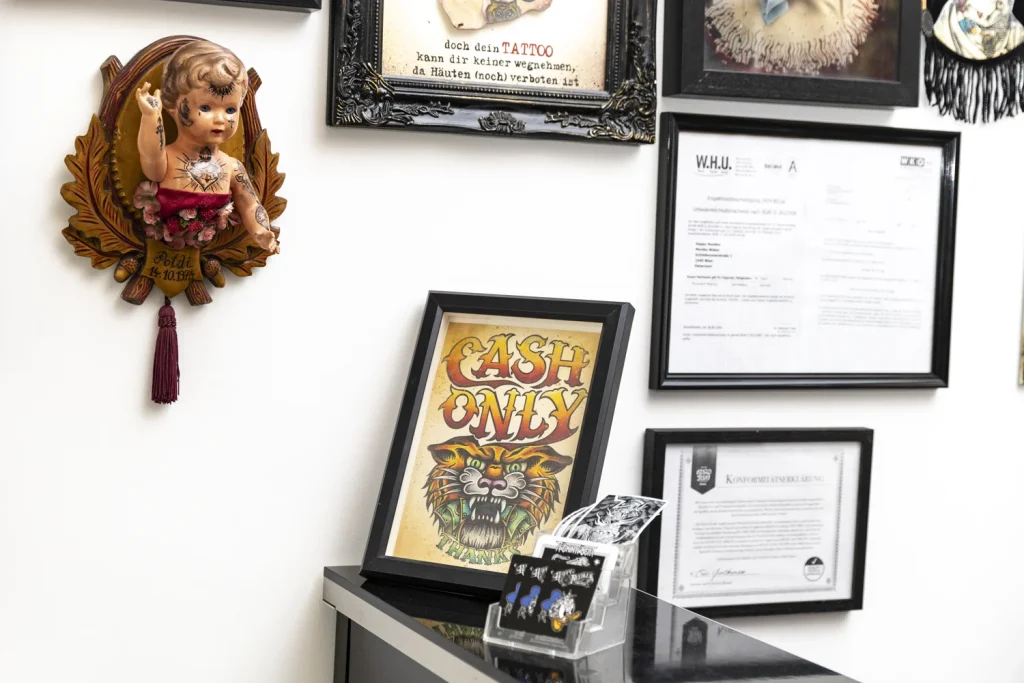 Das Bild zeigt eine Wanddekoration im Tattoo Studio Wien Happy Needles mit mehreren gerahmten Bildern und einem Puppenkopf mit Tattoos auf einem holzgeschnitzten Schild. Das Schild ist mit Blumen und einer Quaste verziert, die anderen Rahmen zeigen verschiedene Bilder und Zertifikate.